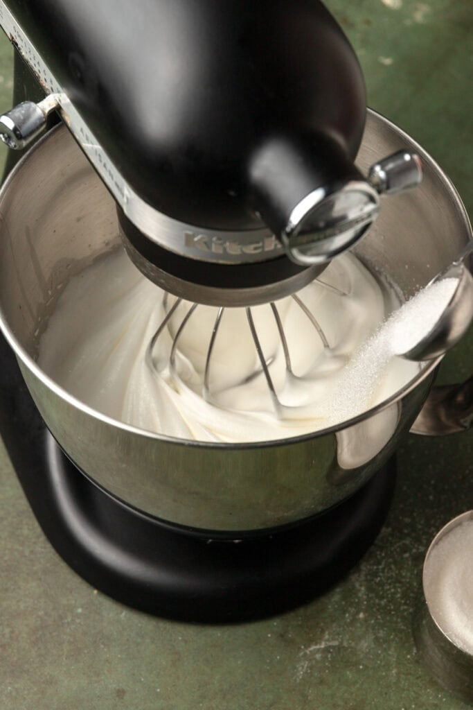 A stand mixer bowl with egg whites adding sugar to it.