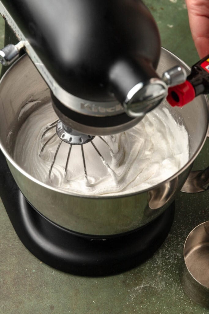 A stand mixer bowl with egg whites adding almond extract to it.