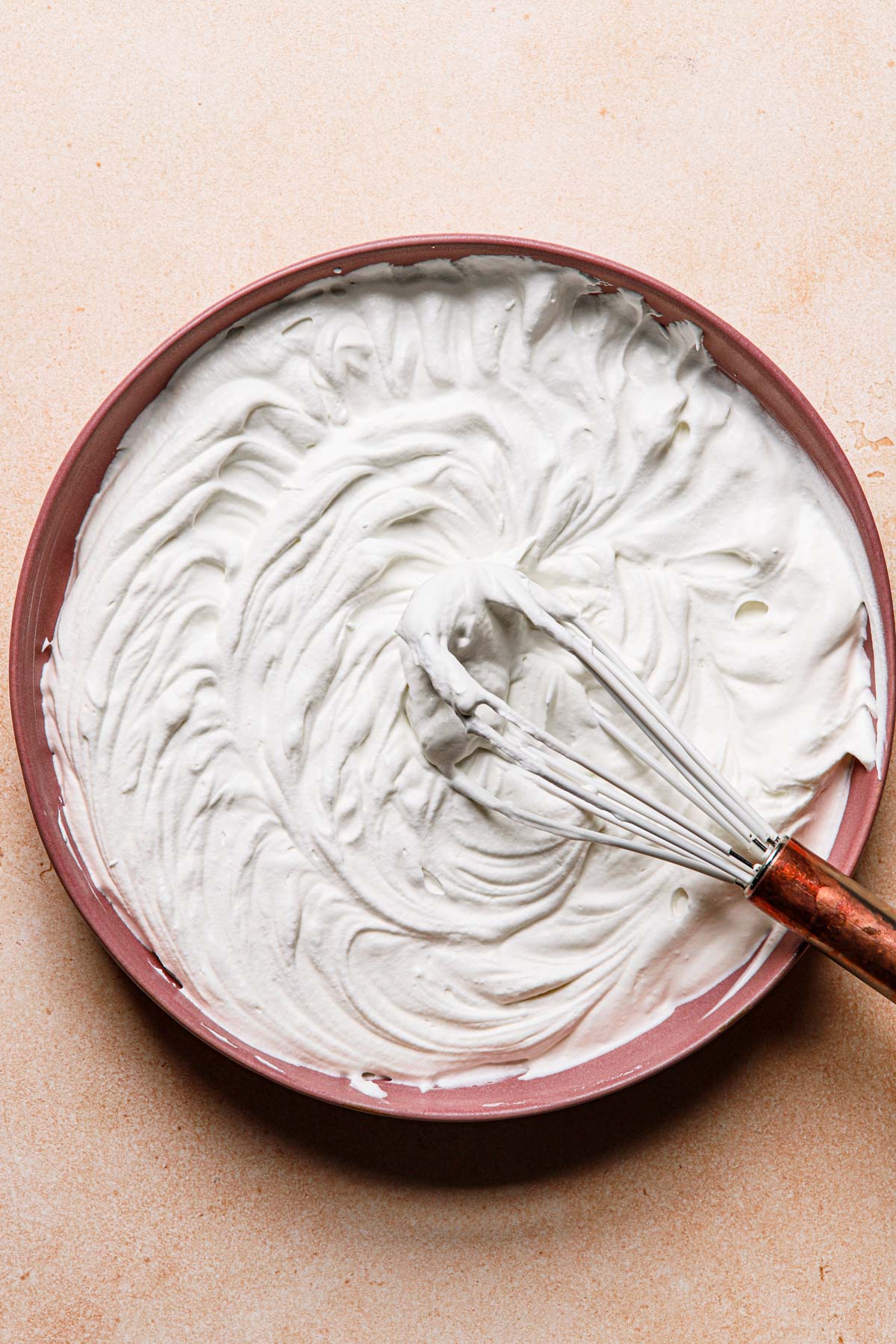 Whipped cream in a pink bowl and a hand whisk.