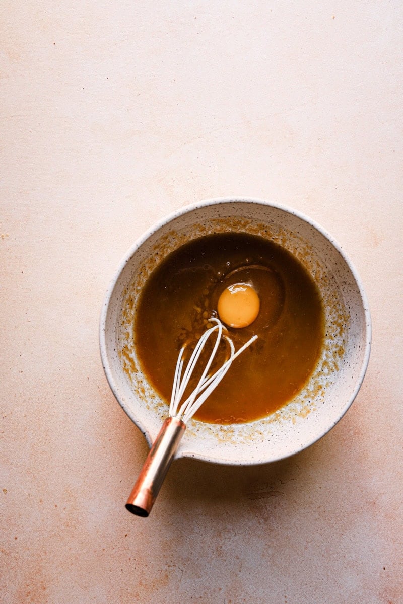 A bowl with melted butter, sugar, and and an egg. A whisk in the bowl.