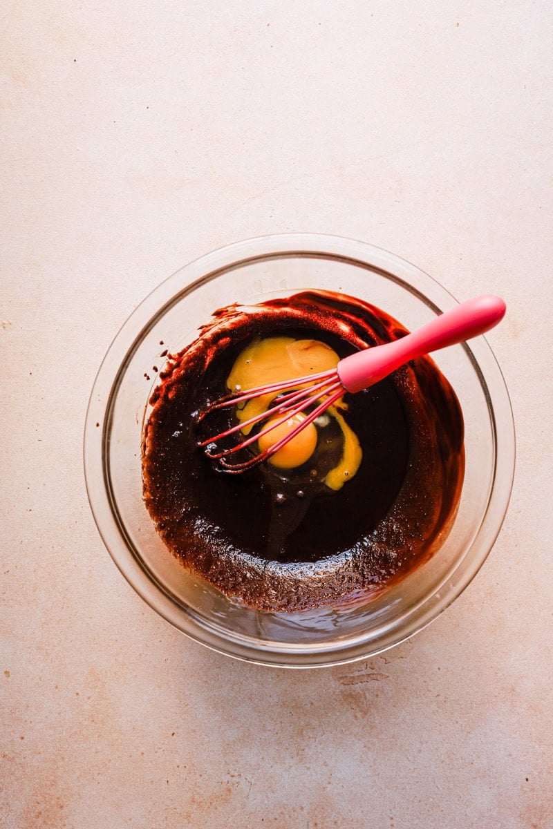 A bowl with melted chocolate, eggs, and a hand whisk.