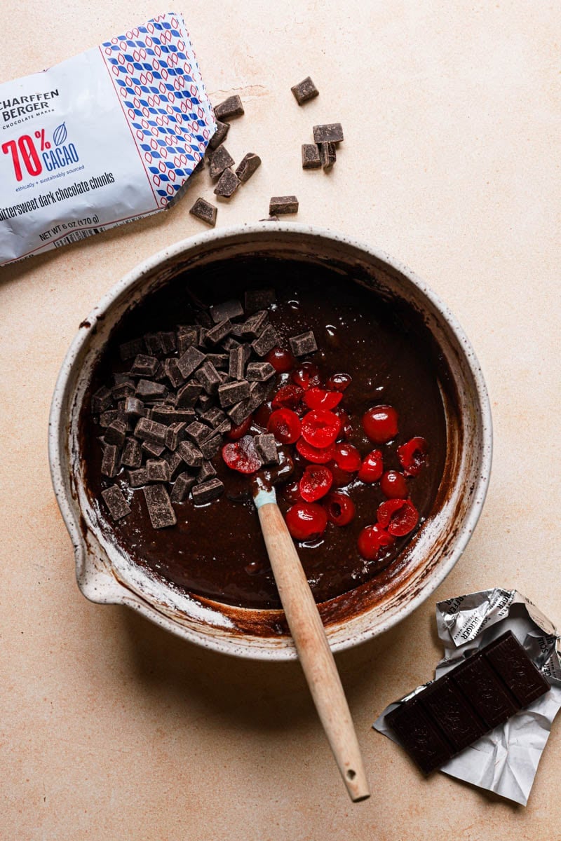A bowl with brownie mixture and a spatula folding in chocolate chips and chopped maraschinos cherries.