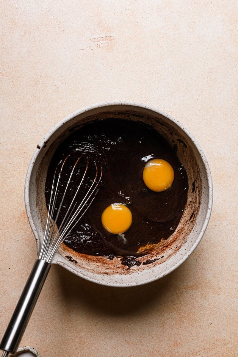 A bowl with chocolate mixture, eggs and a whisk.