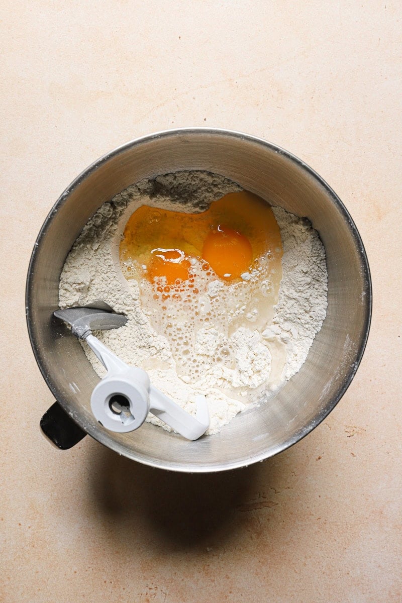 A kitchen mixer bowl with flour, eggs and the paddle attachment.