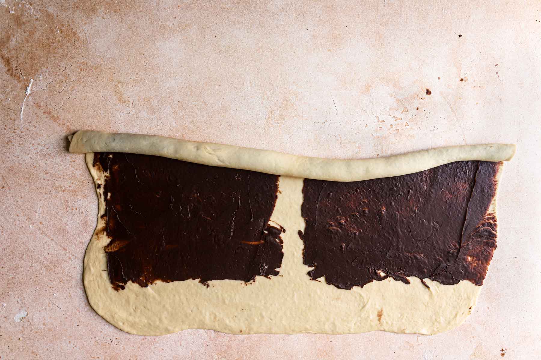 A rectangle dough topped with chocolate filling rolled from he top half way towards the center.
