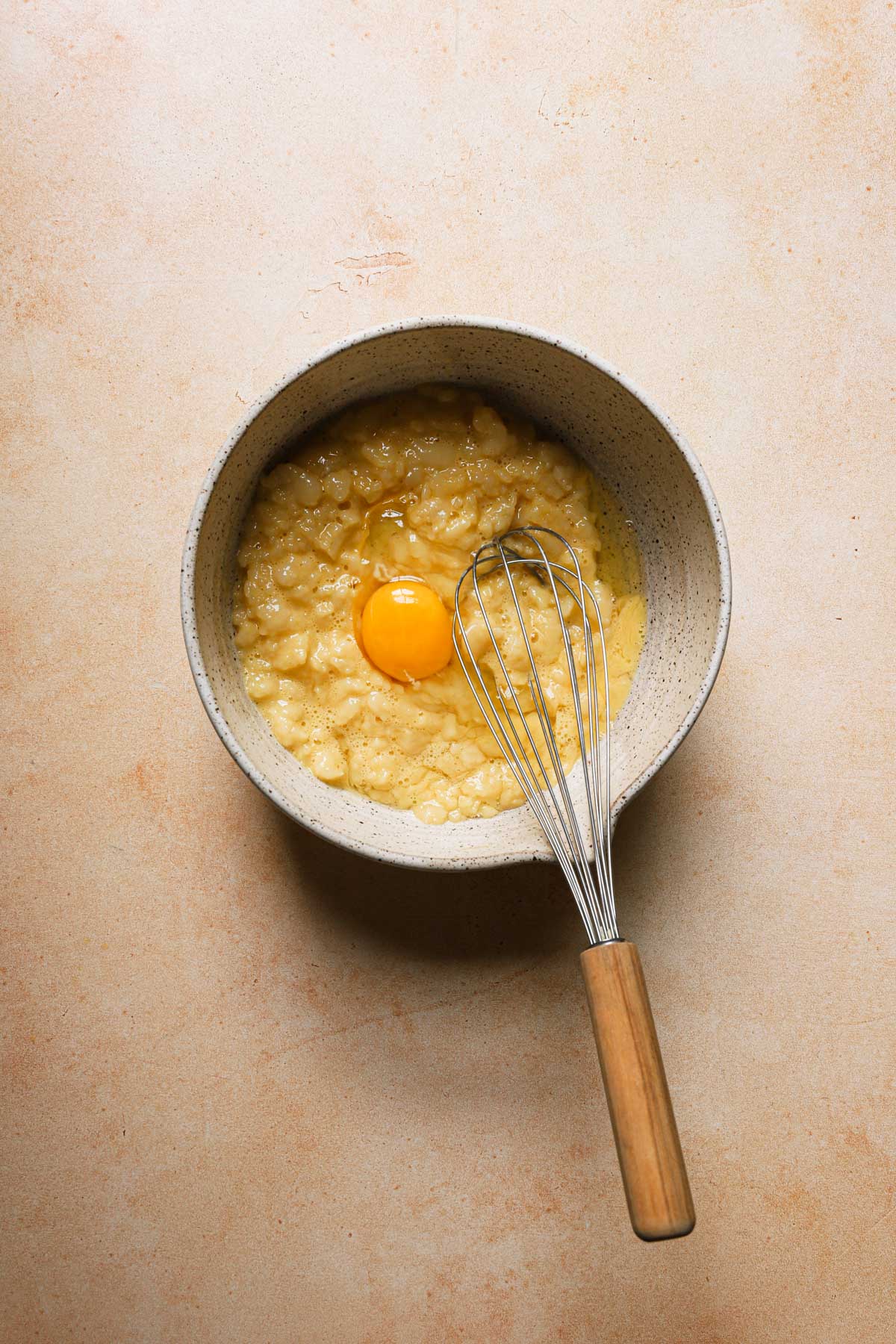 A large bowl with a cream puffs mixture and an egg, a whisk in the bowl.