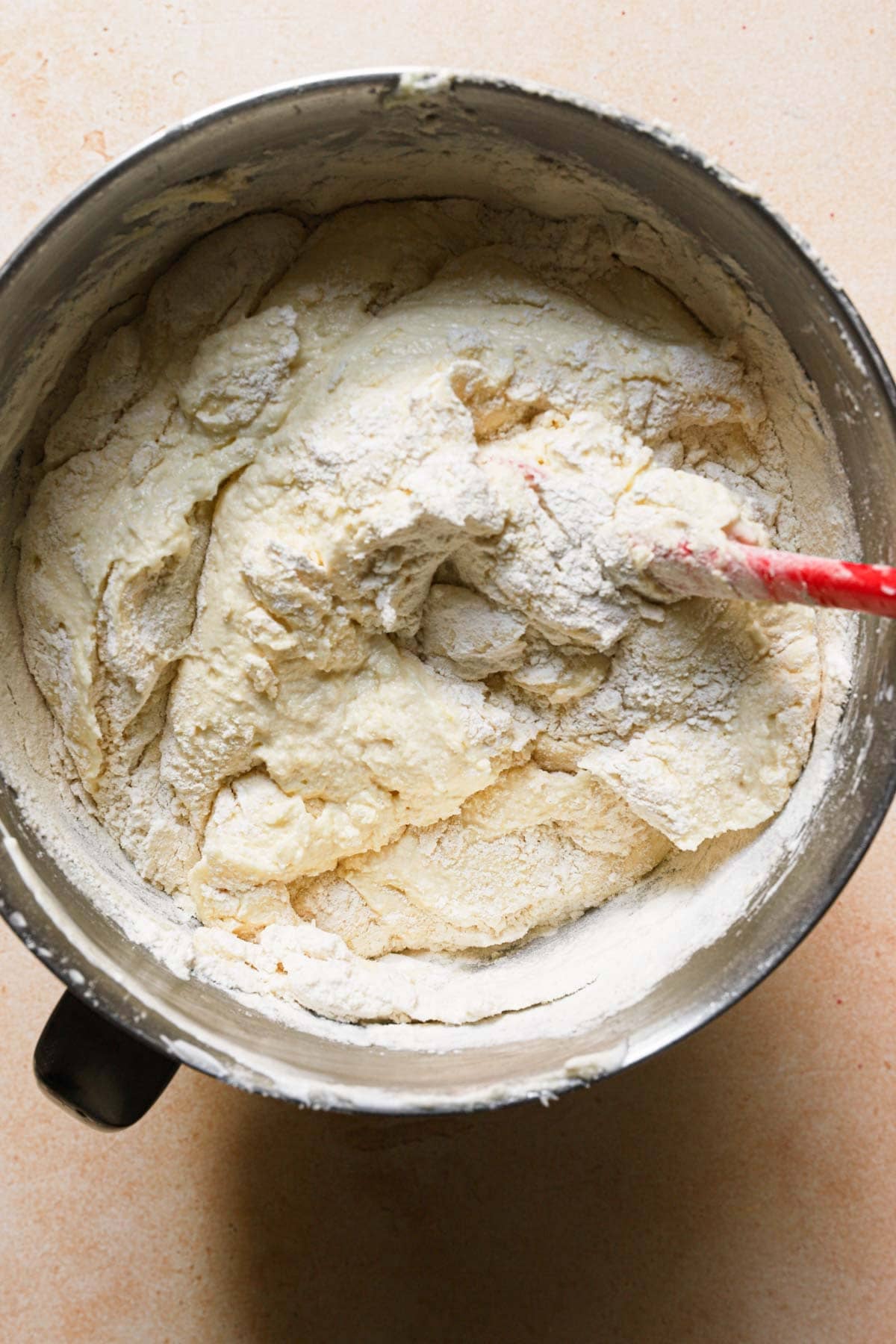 A mixer bowl with a cake batter, dry flour and a rubber spatuala.