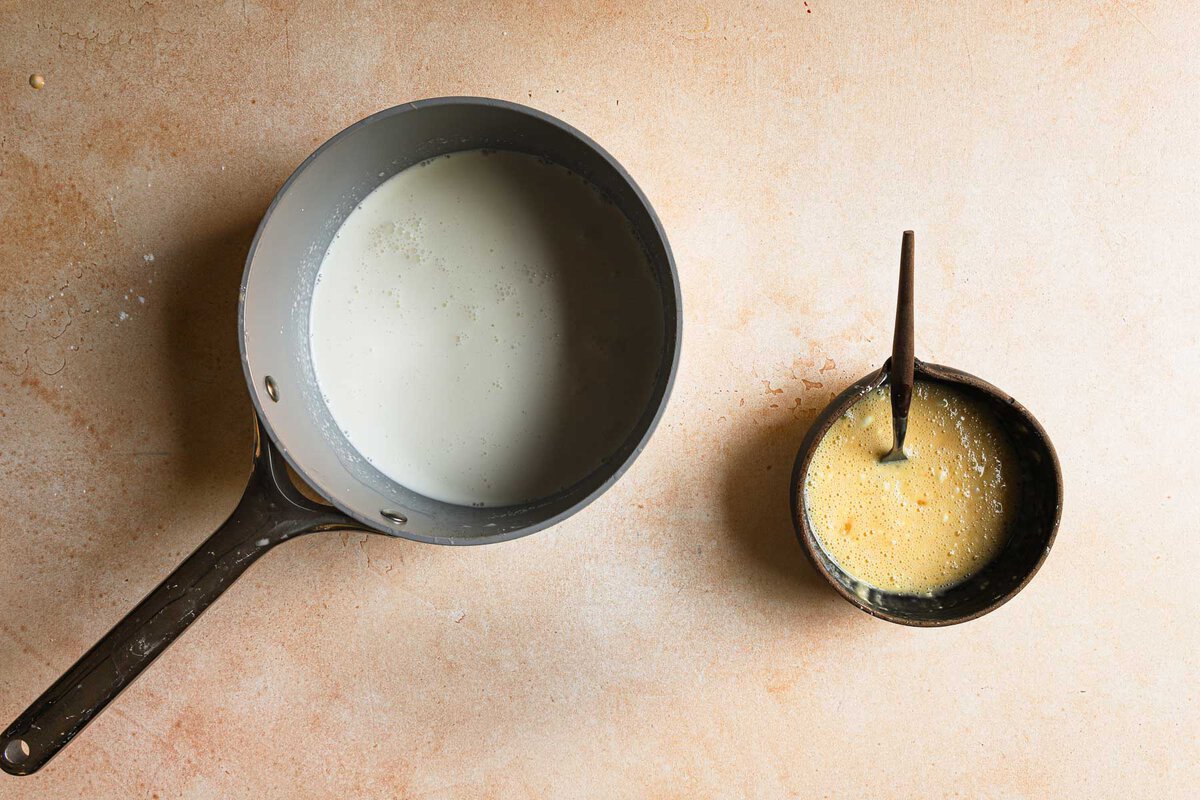 A sauce pan with milk next to a bowl with beaten eggs and cornstarch and a fork.