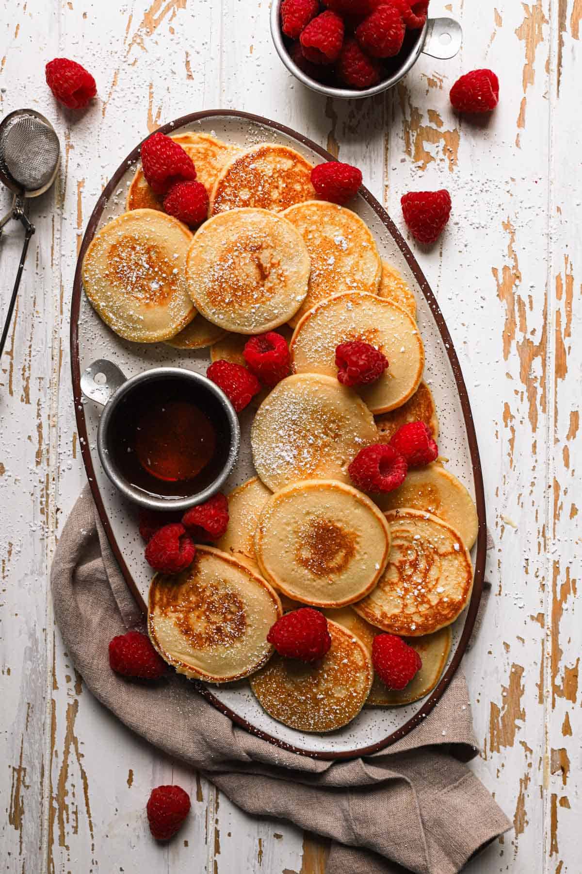 an oval plate with small pan cakes, raspberries and maple syrup