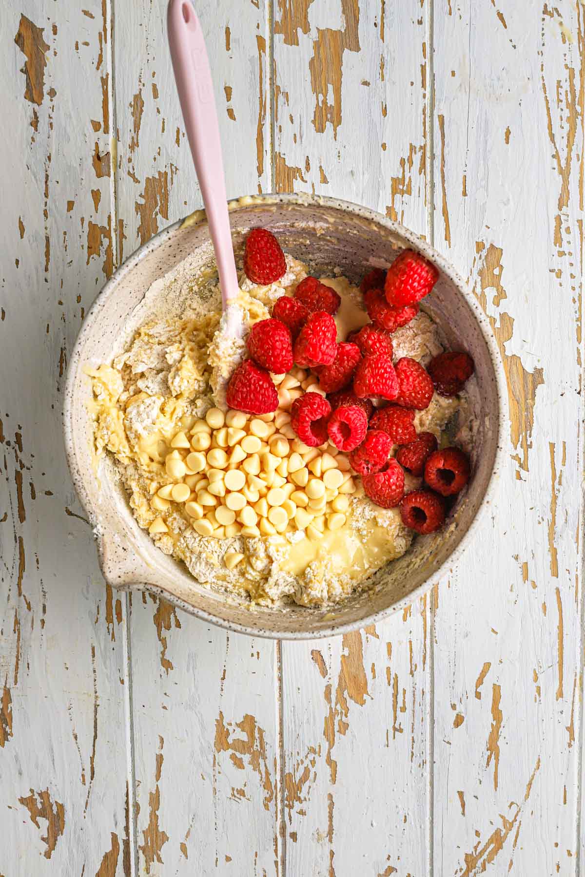 A large bowl with a spatula and muffins batter plus fresh raspberries and white chocolate chips.