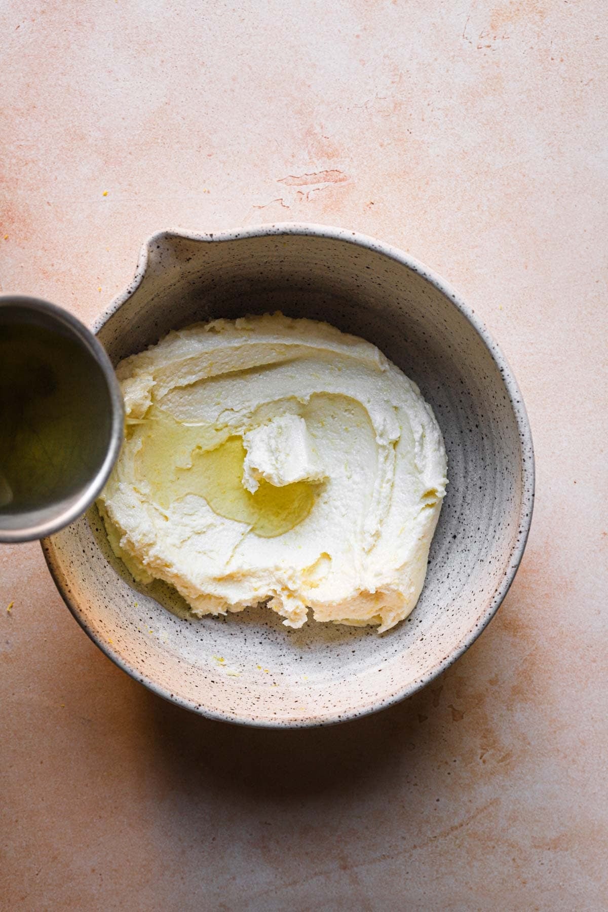 A cup pouring olive oil into a creamed butter and sugar in a bowl.
