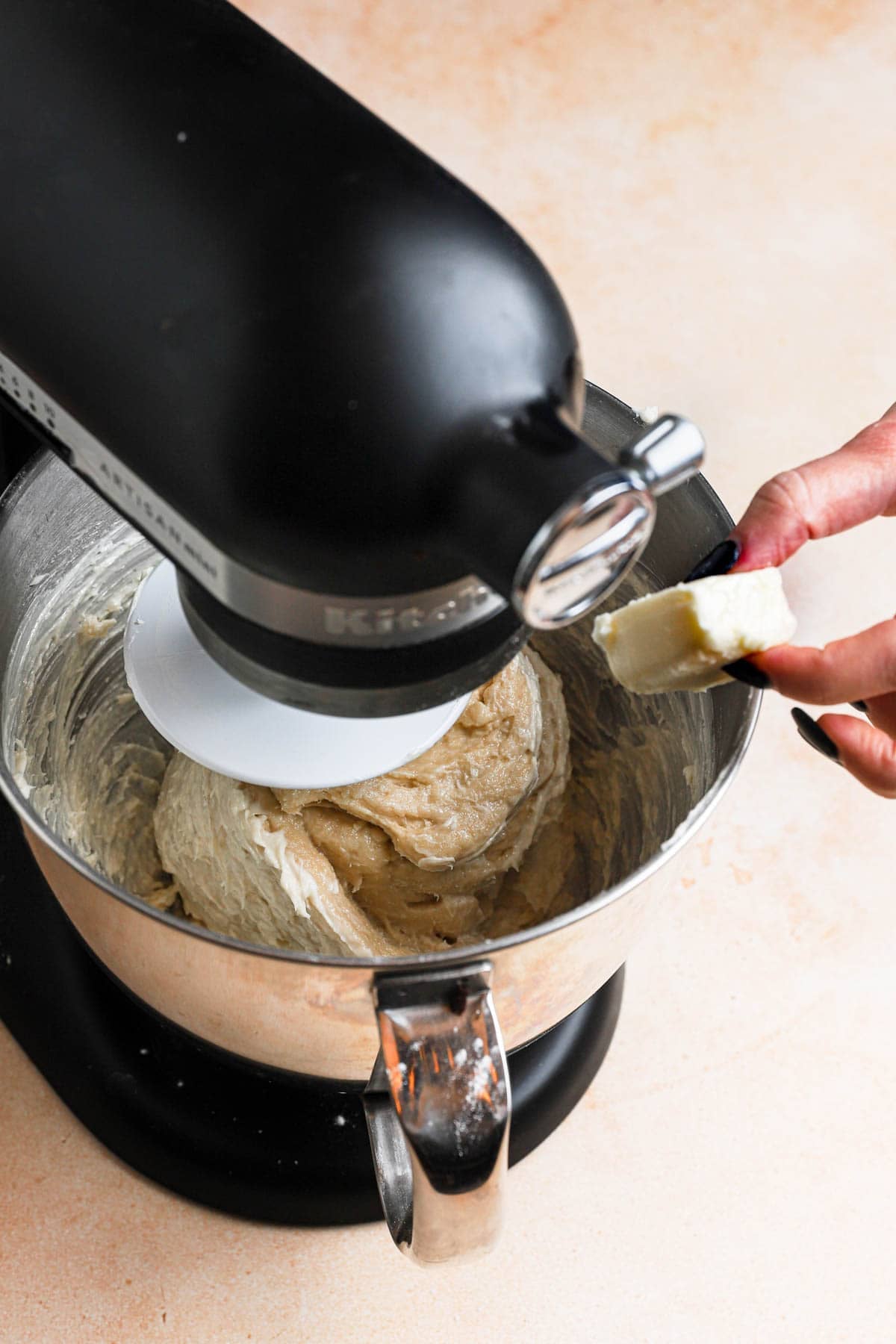 Adding butter to a bowl of a stand mixer fitted with the dough hook and dough inside.