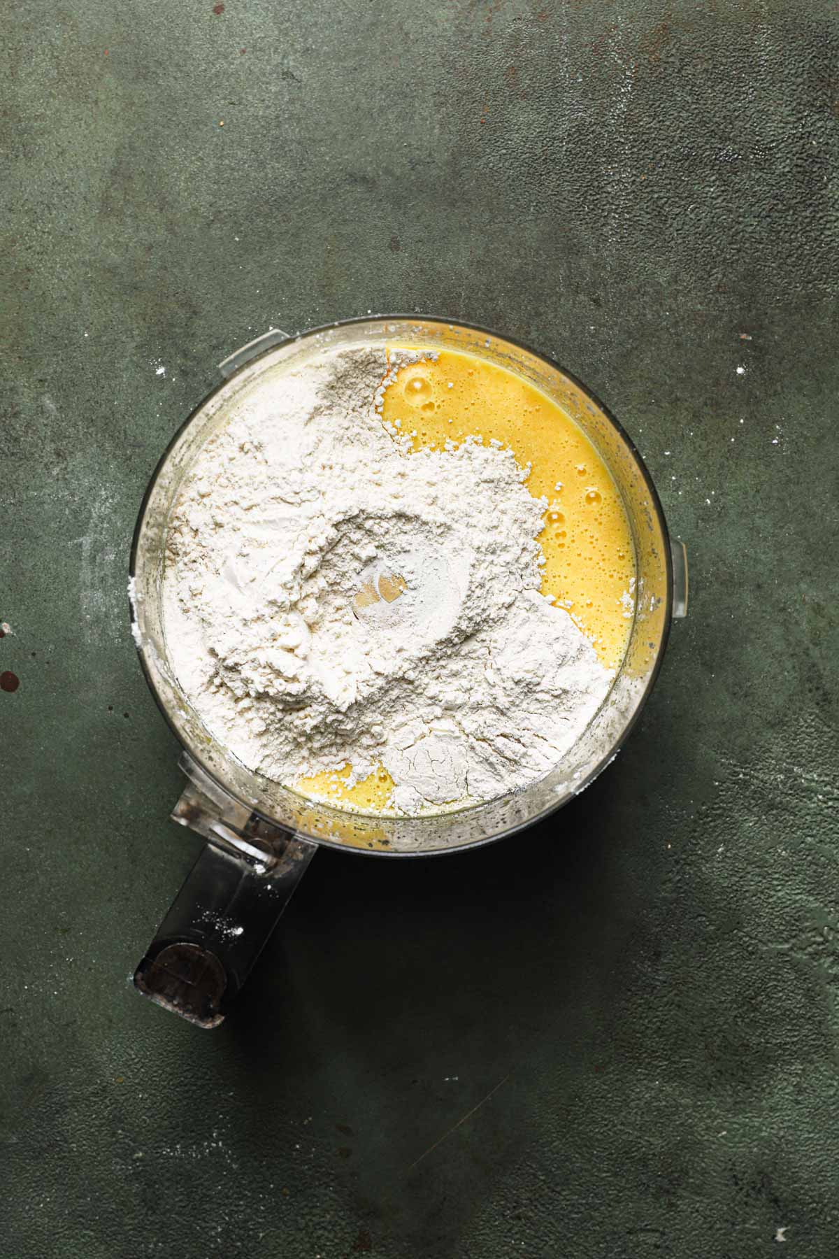 A food processor bowl with orange cake batter and flour.