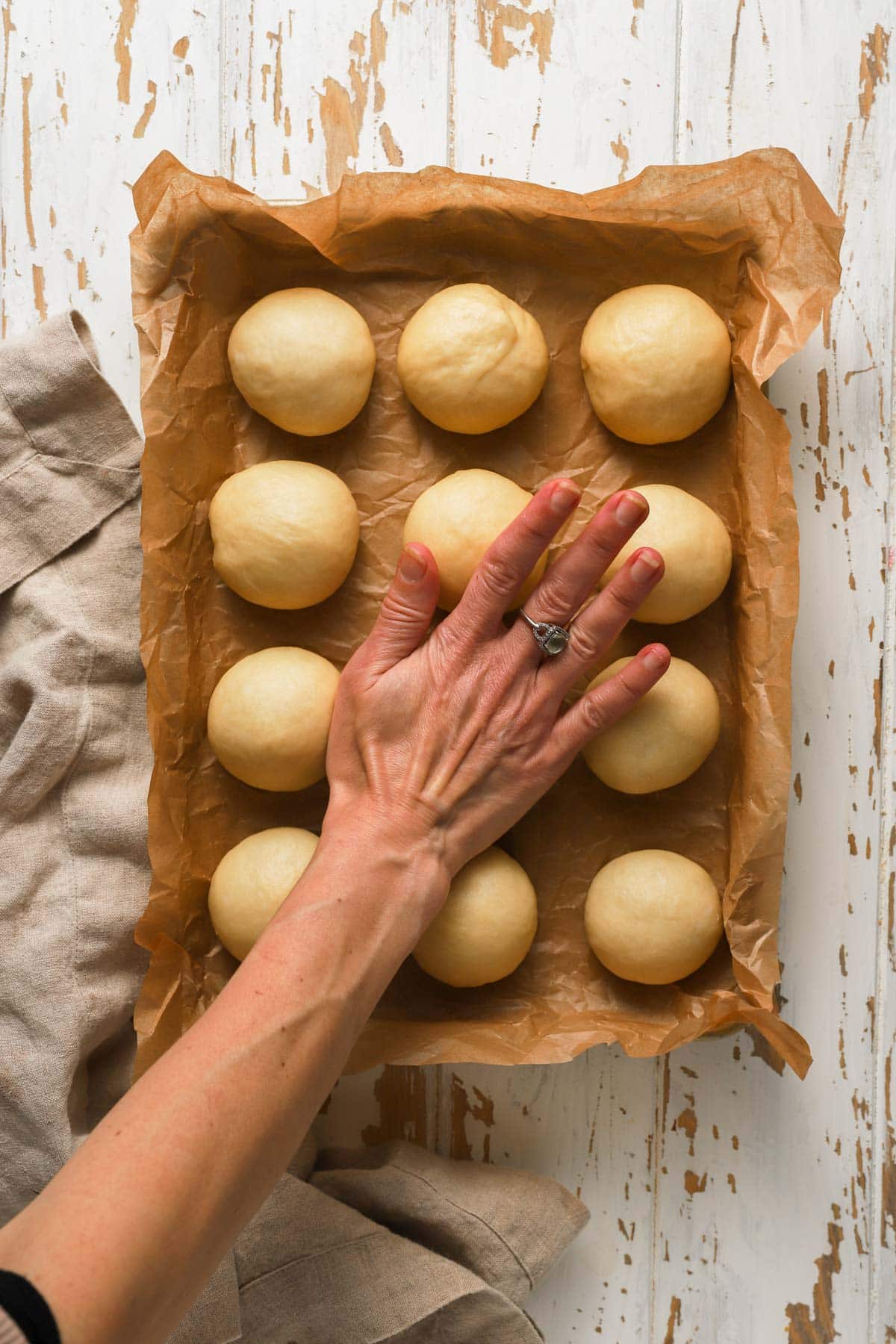 A hand pressing dinner rolls over a pan with parchment paper.