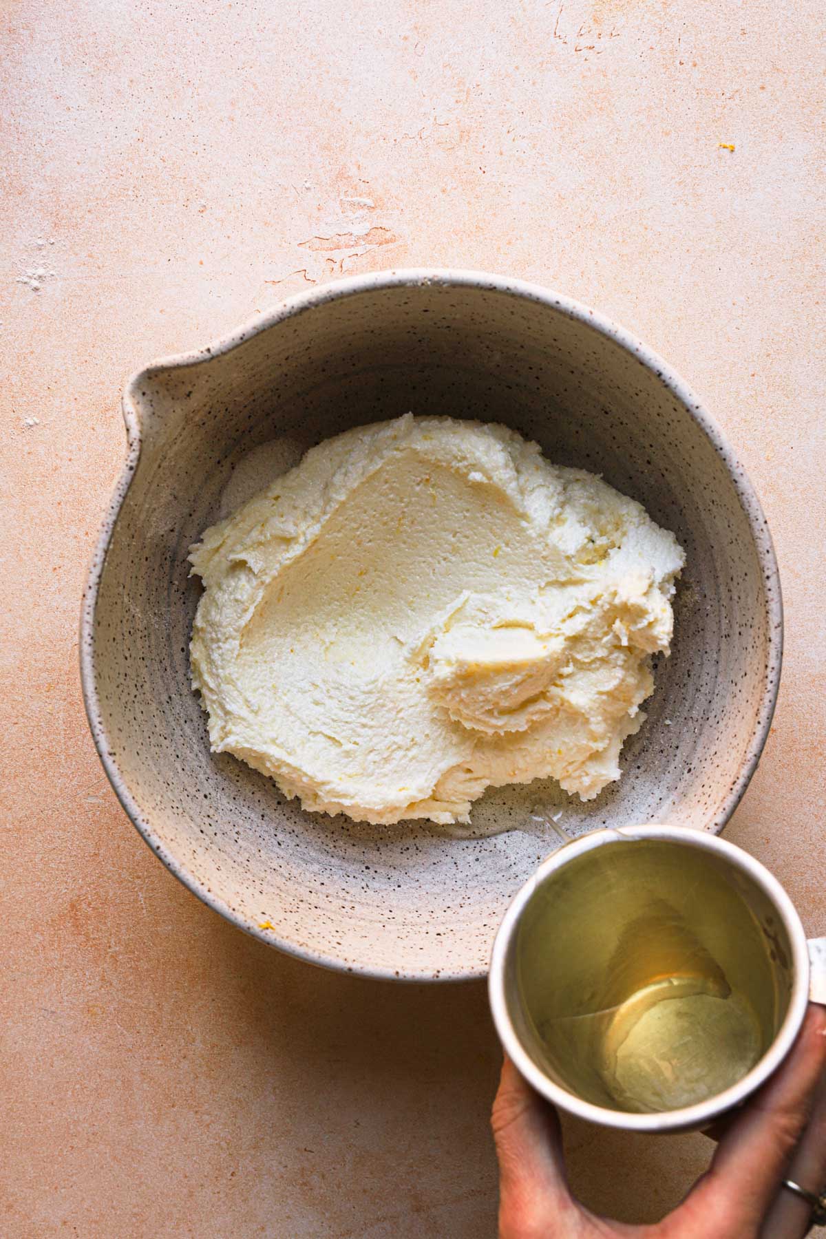 A hand pouring olive oil into a bowl with creamed sugar and butter.