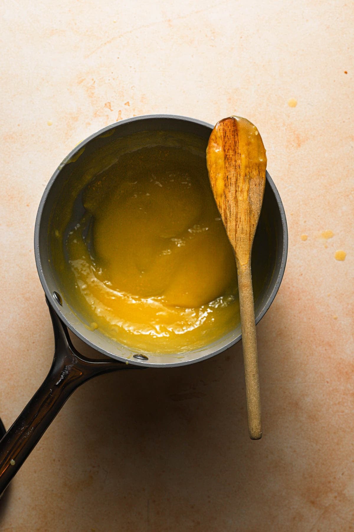 Thickened lemon curd n a sauce pan with a wooden spoon on top.