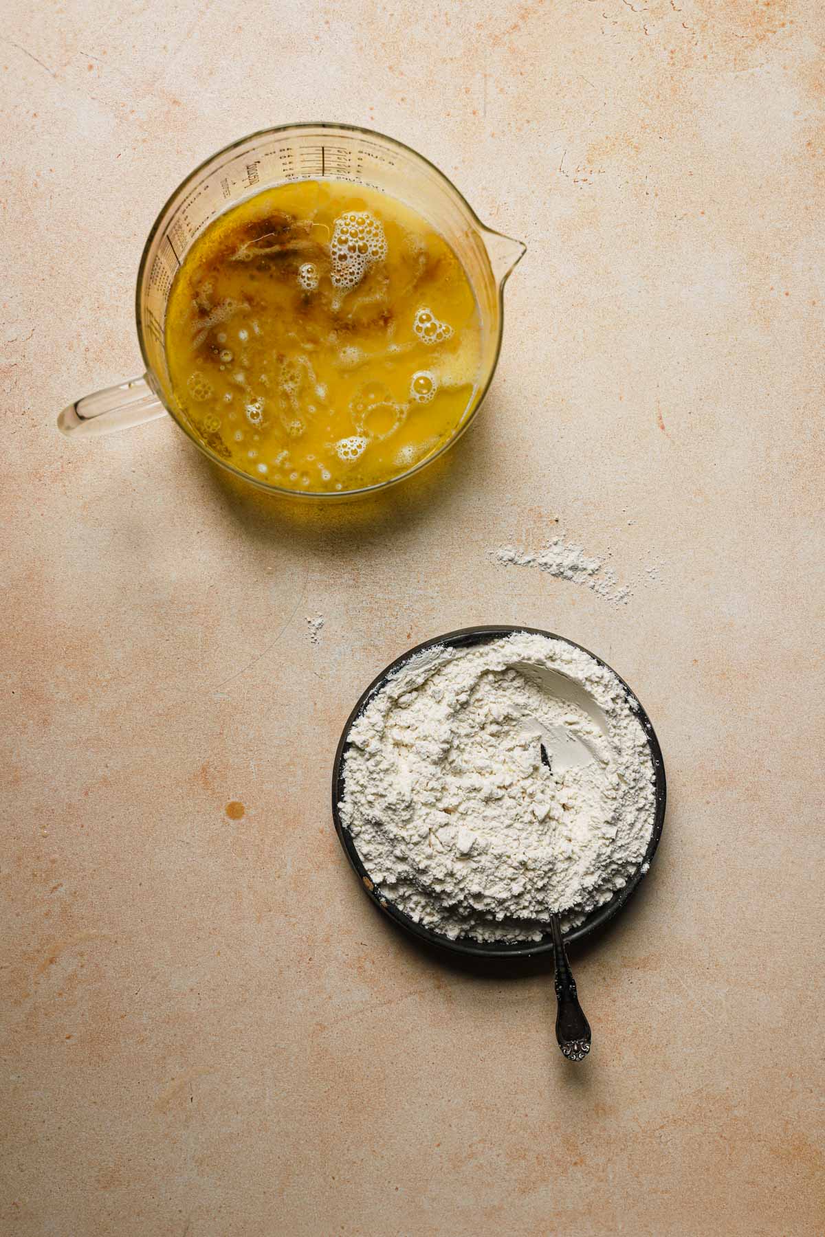 A measuring cups with eggs mixture and a bowl with flour mixture.