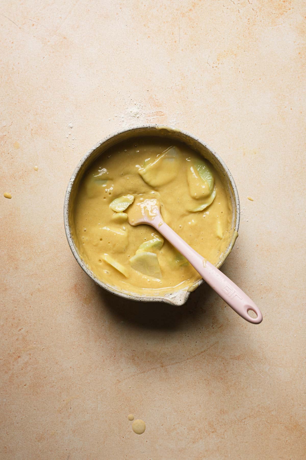 A bowl with cake batter and sliced apples with a spatula.