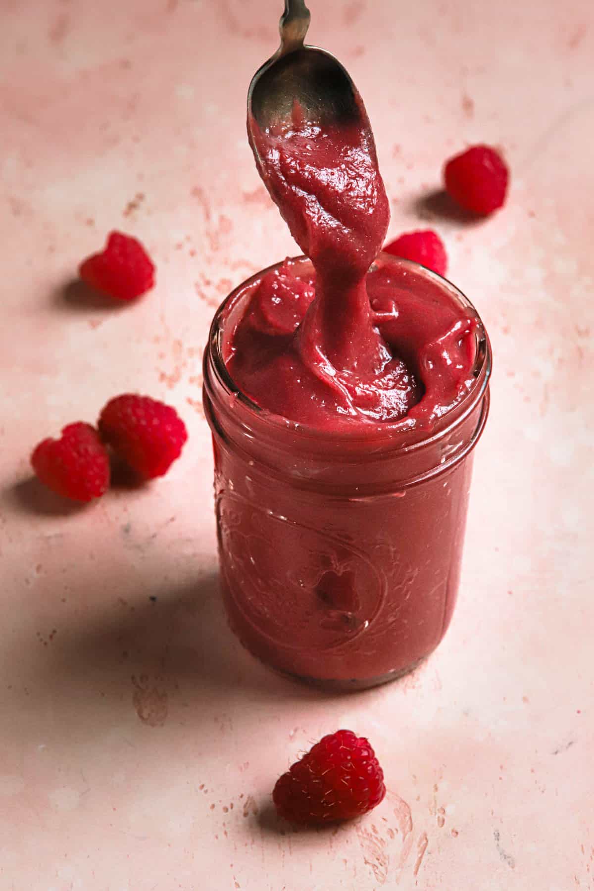 raspberry curd in a mason jar and a spoon with raspberries around it.