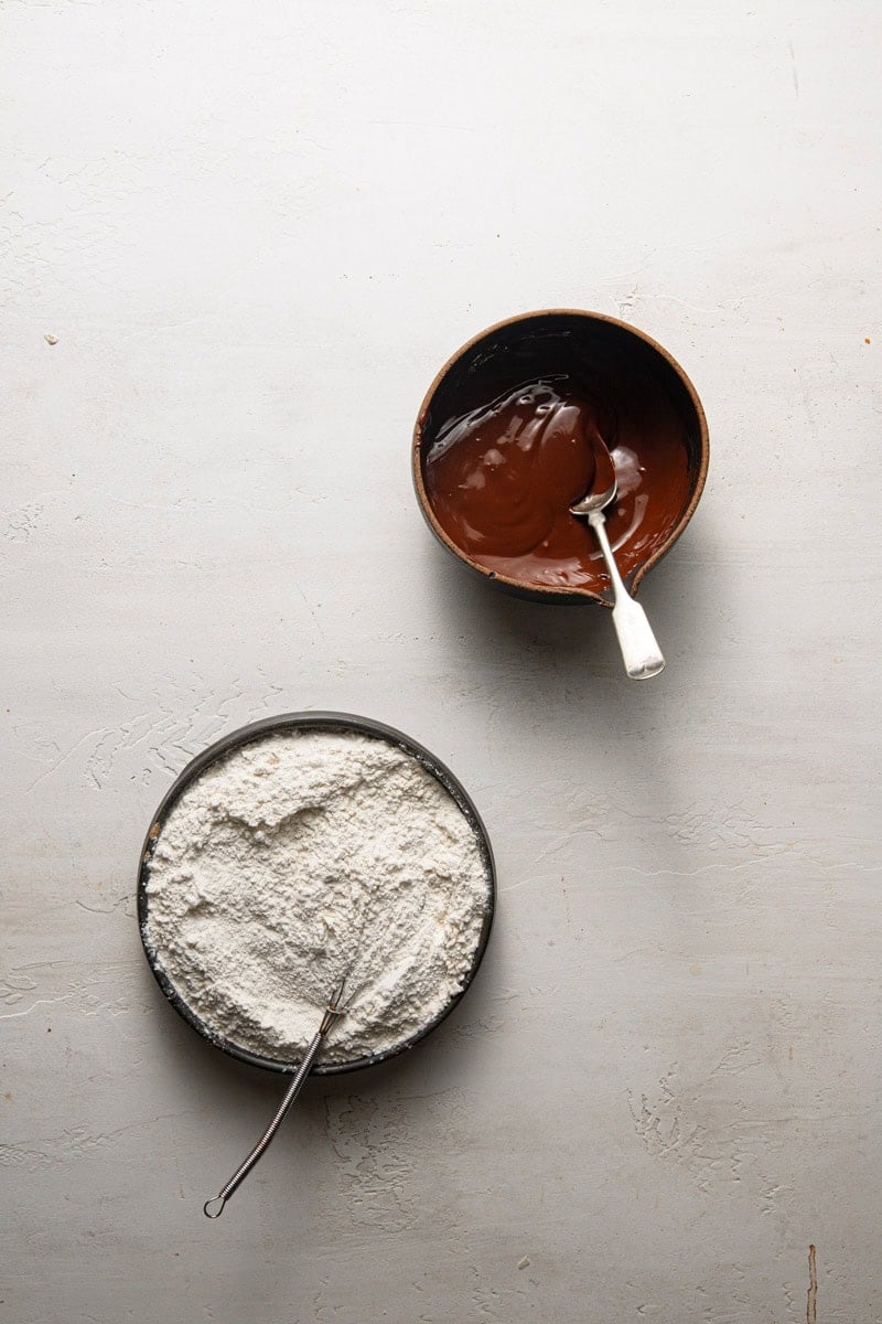 A bowl with melted chocolate next to a bowl with flour.
