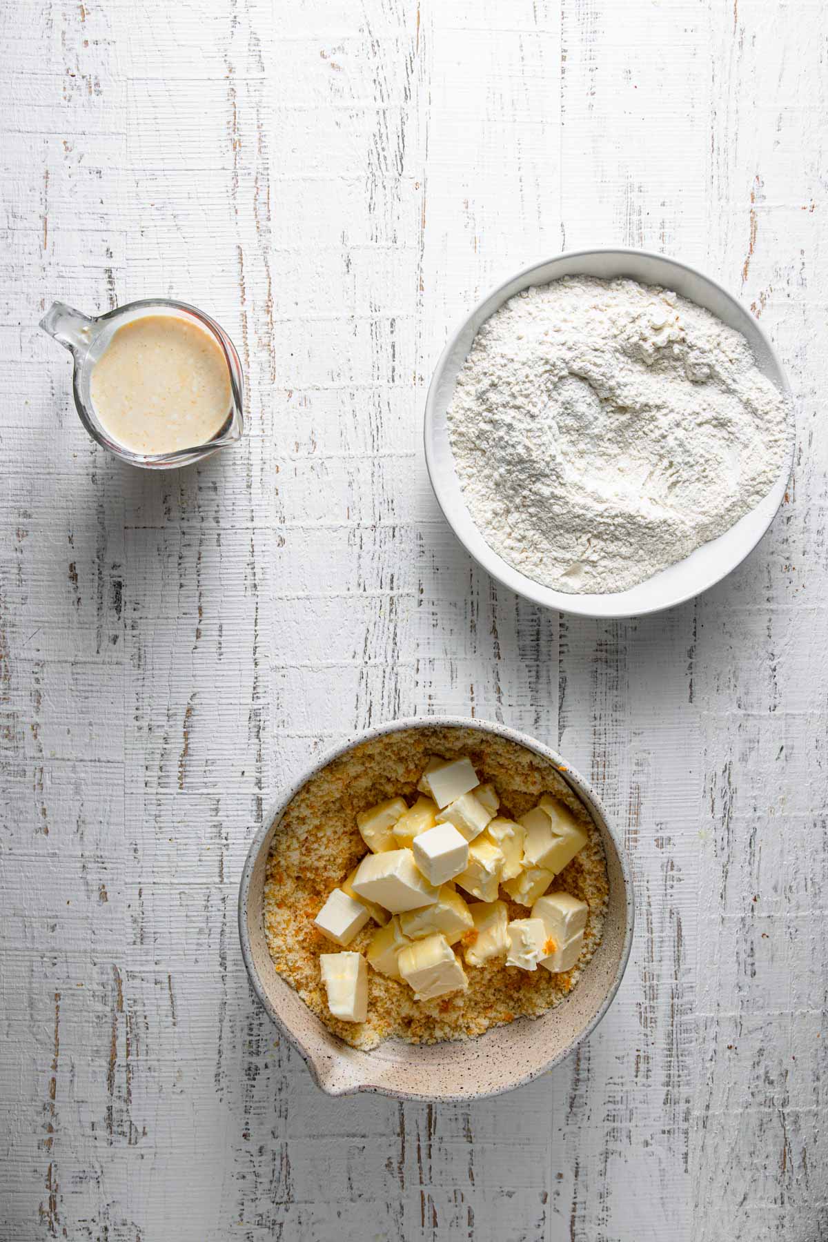 A bowl with flour mixture, a bowl with orange sugar and butter and a measuring cup with sour cream.
