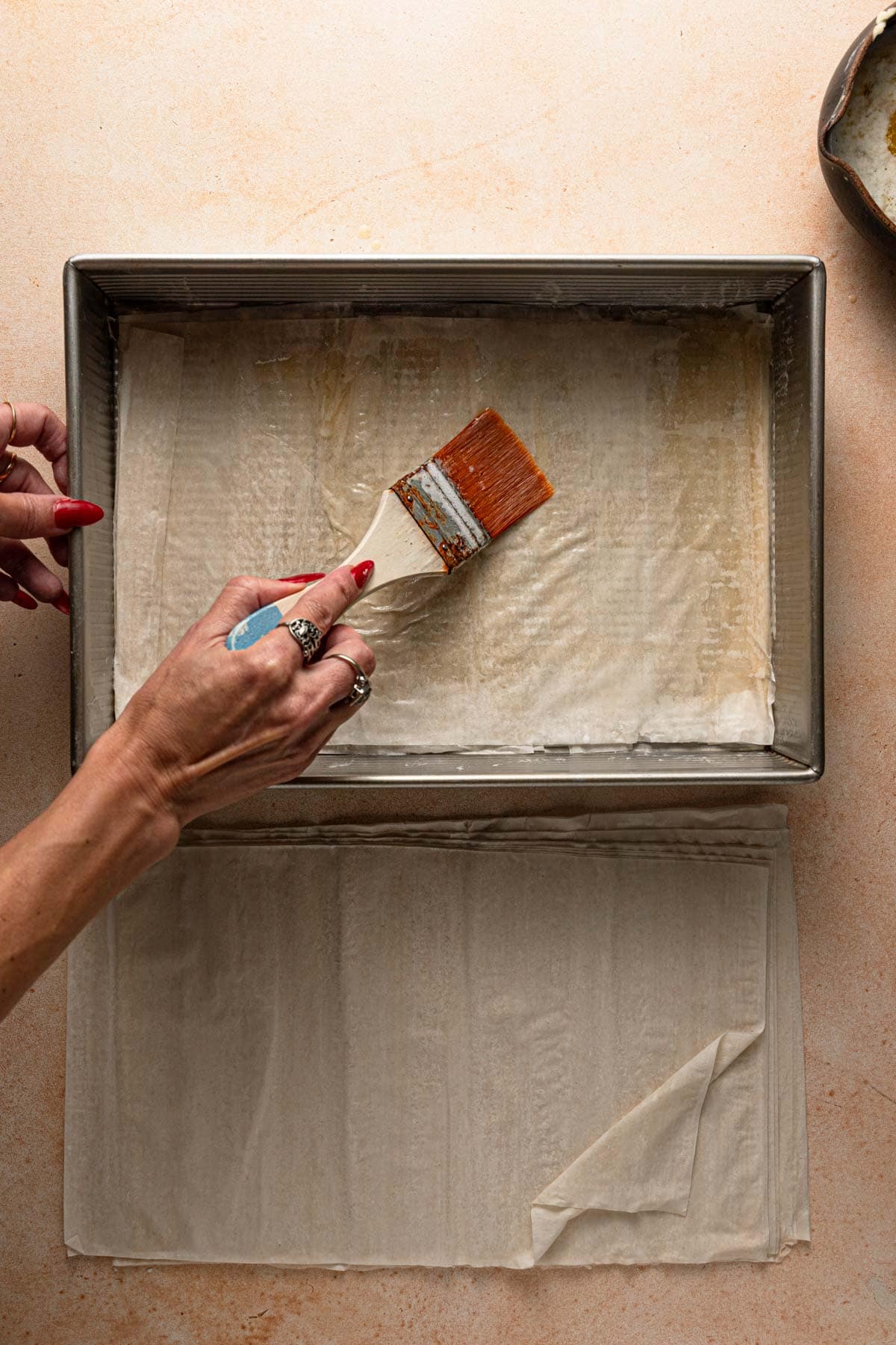 Hands brushing phyllo sheet lined in a pan.