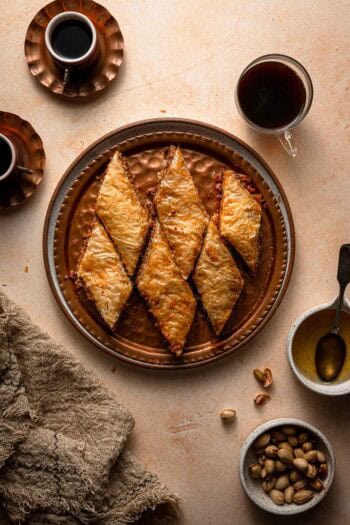 Phillo dough and nuts dessert soaked in honey syrup on a serving plate with coffee cups and pistachios.
