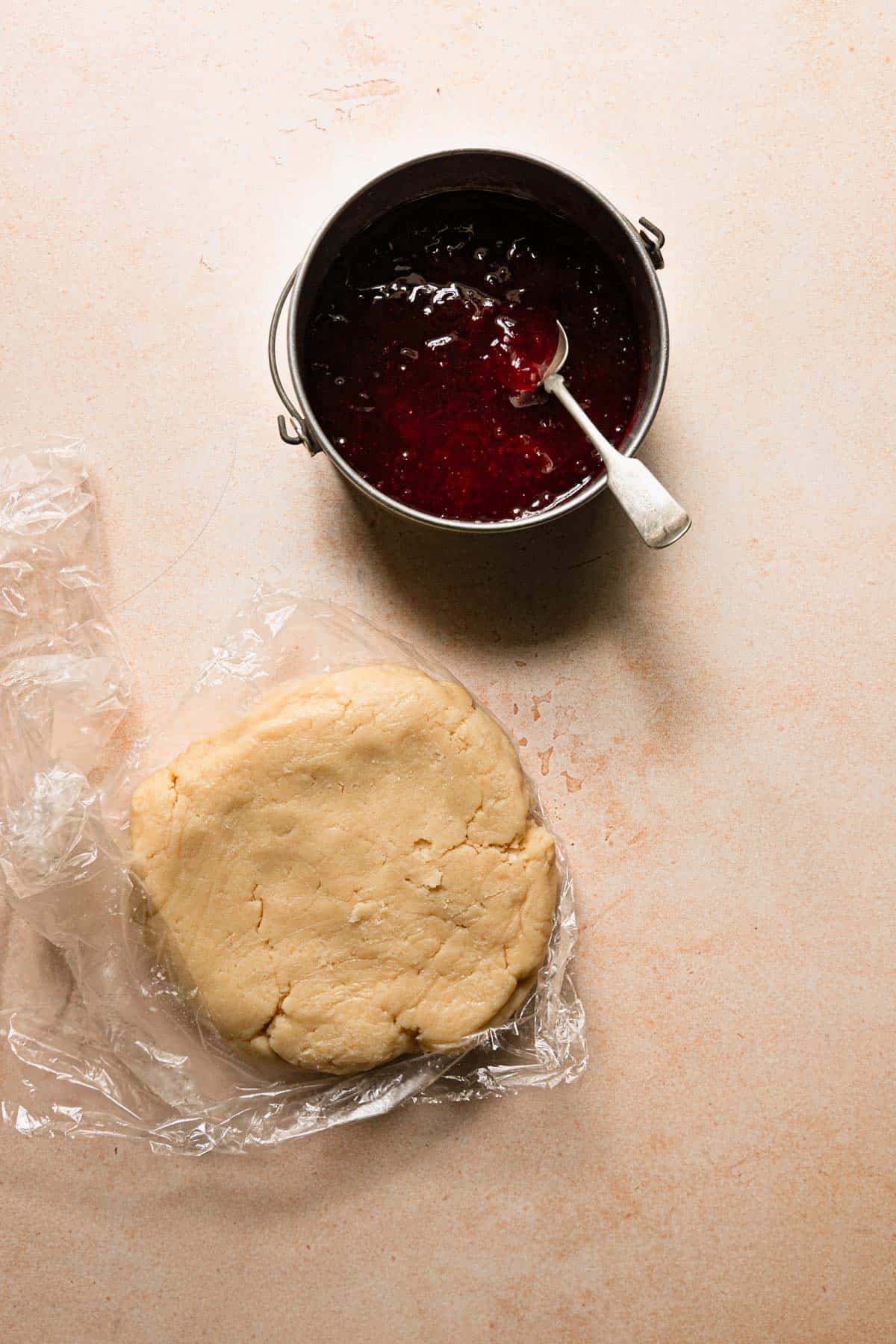 A sauce pan with strawberry preserves and a spoon, next to shortbread dough topped on plastic wrap.