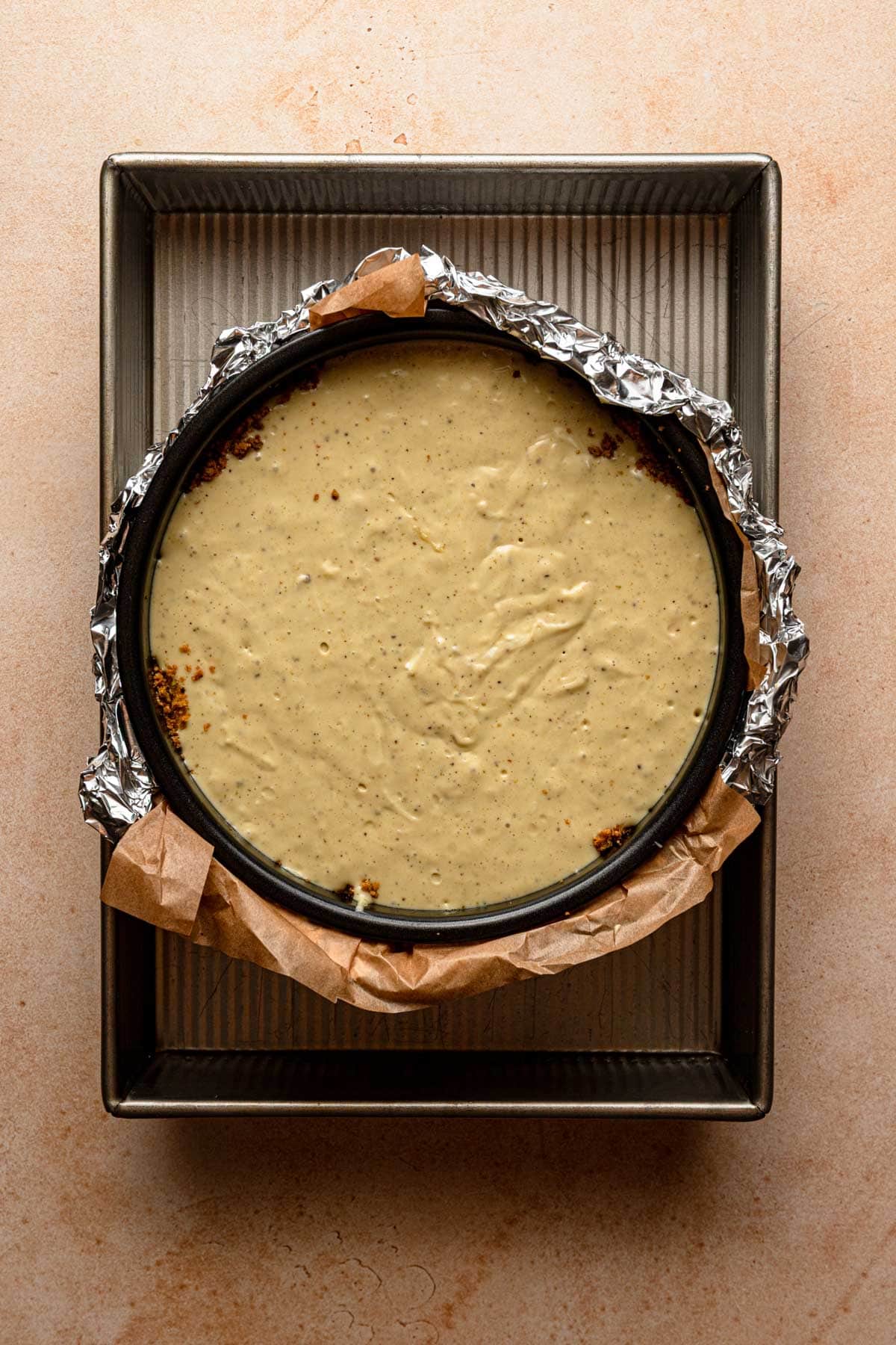 A pan wrapped with aluminum foil and filled with pistachio batter inside a 9x13-inch pan filled with water.