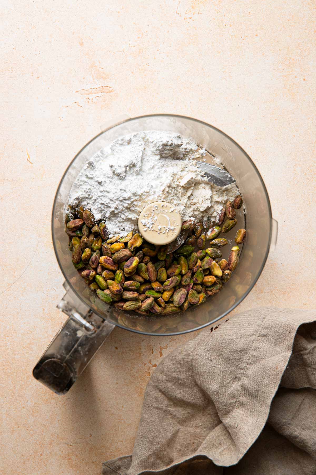 A bowl of a food processor with pistachio and powdered sugar. A napkin next to it.