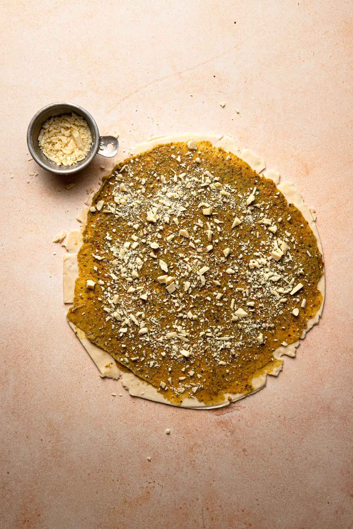 A round rolled dough topped with pistachio cream and chopped white chocolate. A small bowl with chopped chocolate next to it.