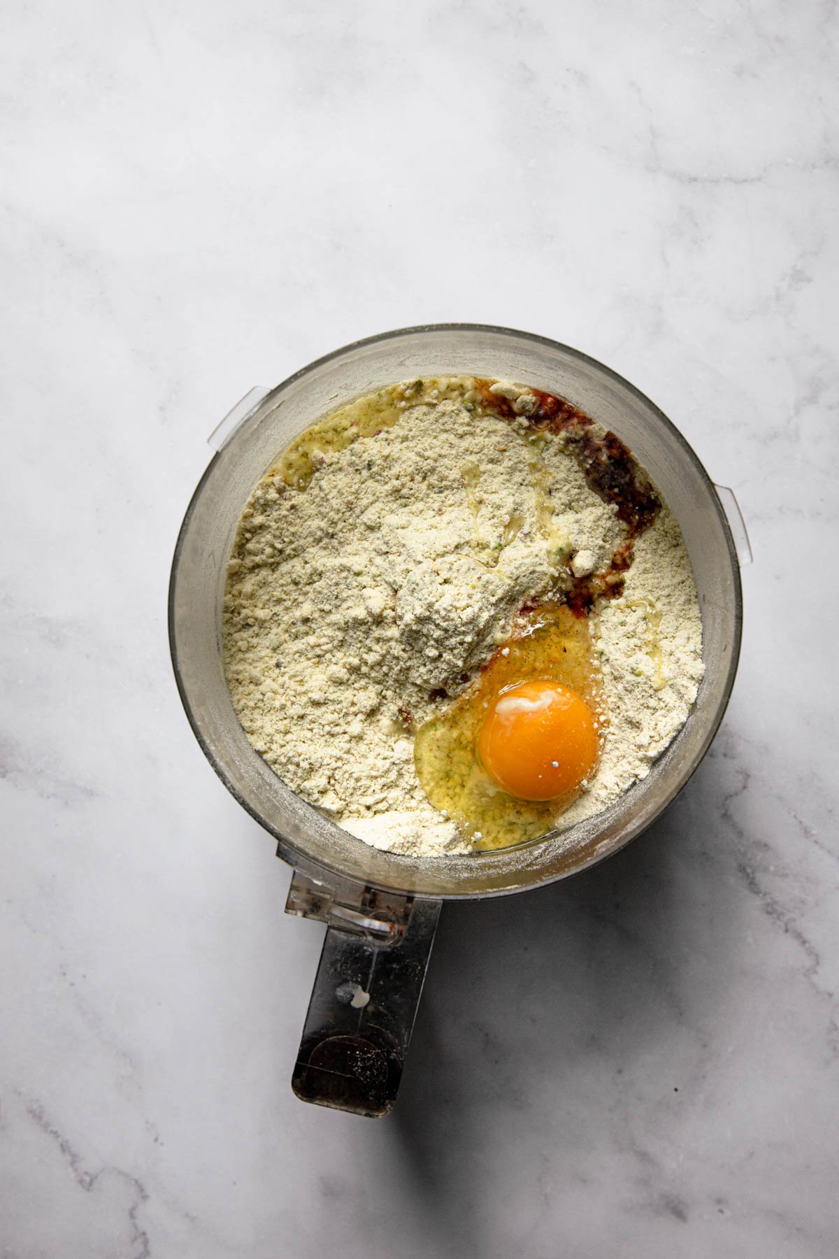 A food processor bowl with flour mixture, egg and vanilla extact.