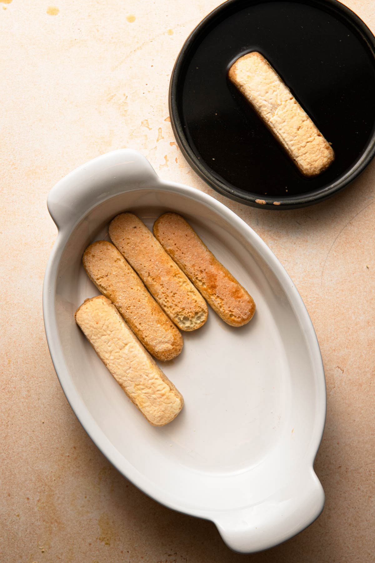A bowl with coffee and a ladyfinger and a serving oval dish with some ladyfingers.