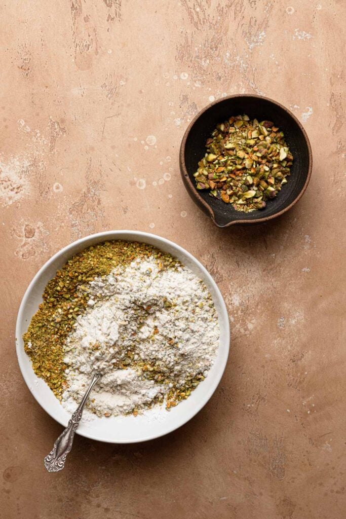 A large bowl with flour and ground pistachios with a fork, next to a smaller bowl with chopped pistachios.
