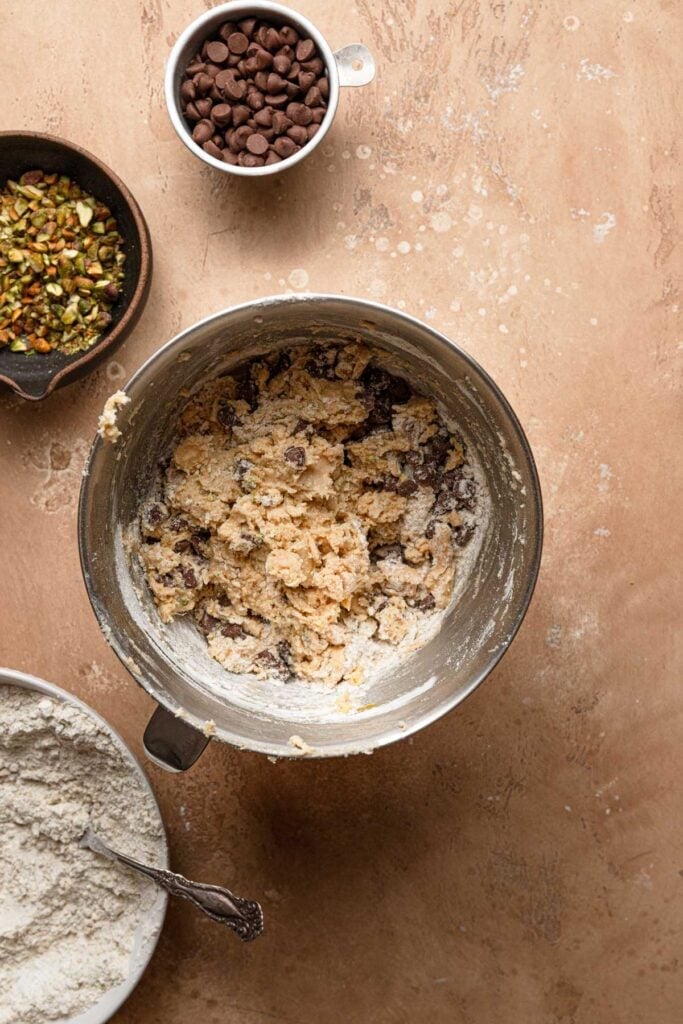 A mixing bowl with cookie dough and next to it bowls with chopped pistachios, chocolate chips and flour.