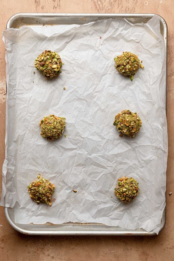 A pan lined with parchment paper and topped with six pistachio chocolate chipe dough balls.