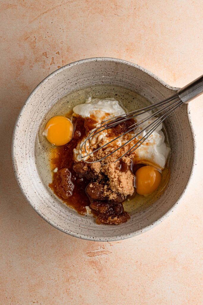 A mixing bowl with vegetable oil, light brown sugar, eggs and sour cream with a whisk.