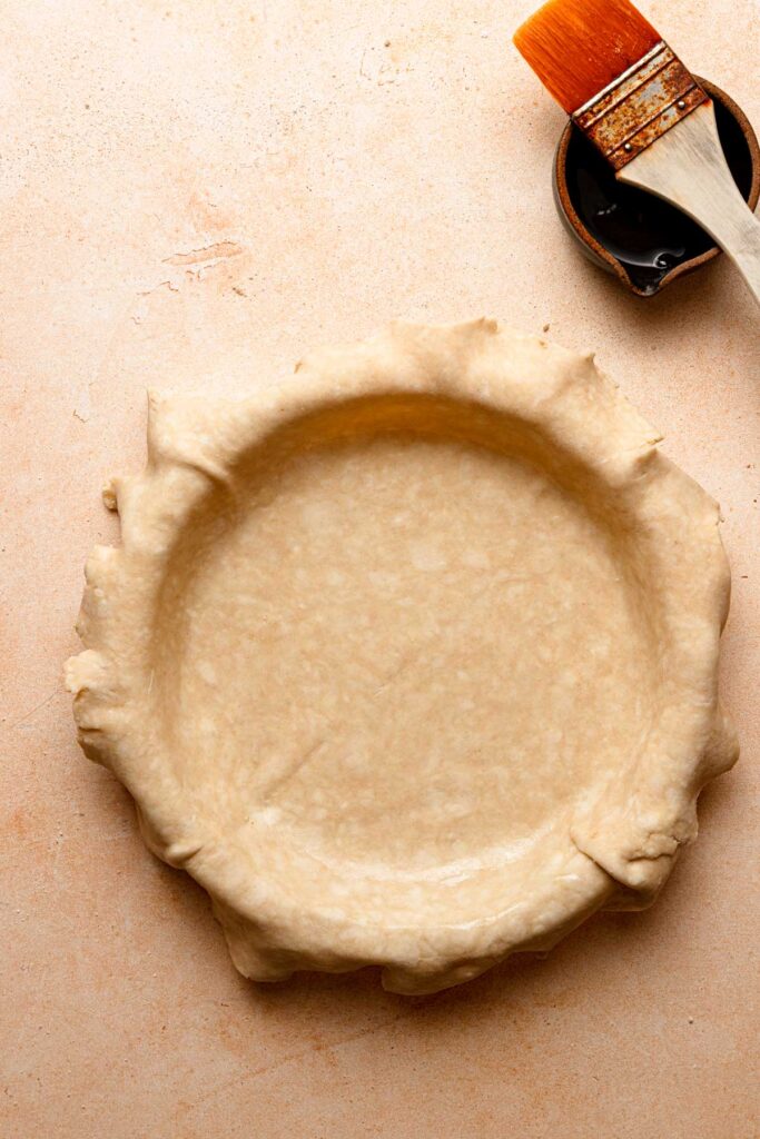 A pie shell topped with pie crust next to a bowl and a pastry brush.