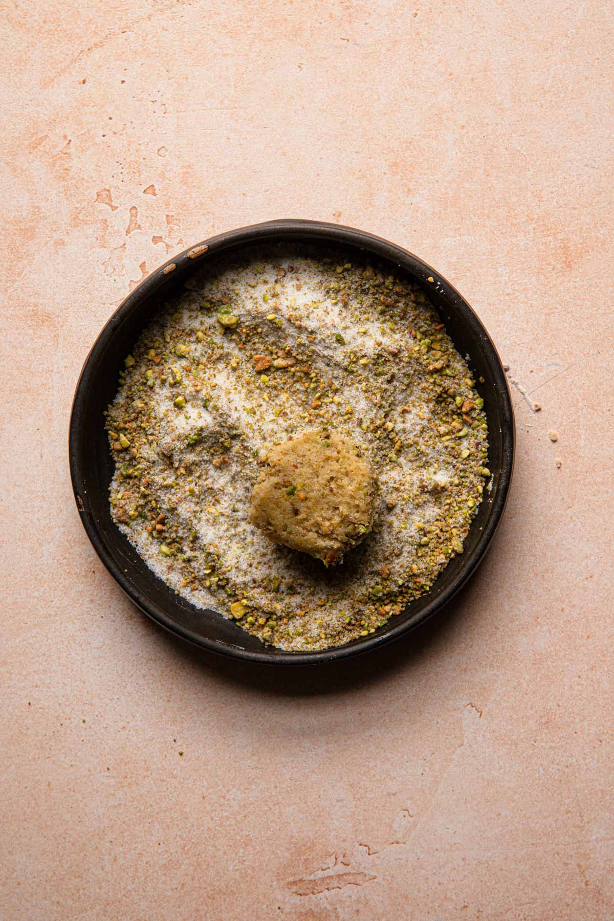A cookie ball in a bowl full of sugar and ground pistachios.