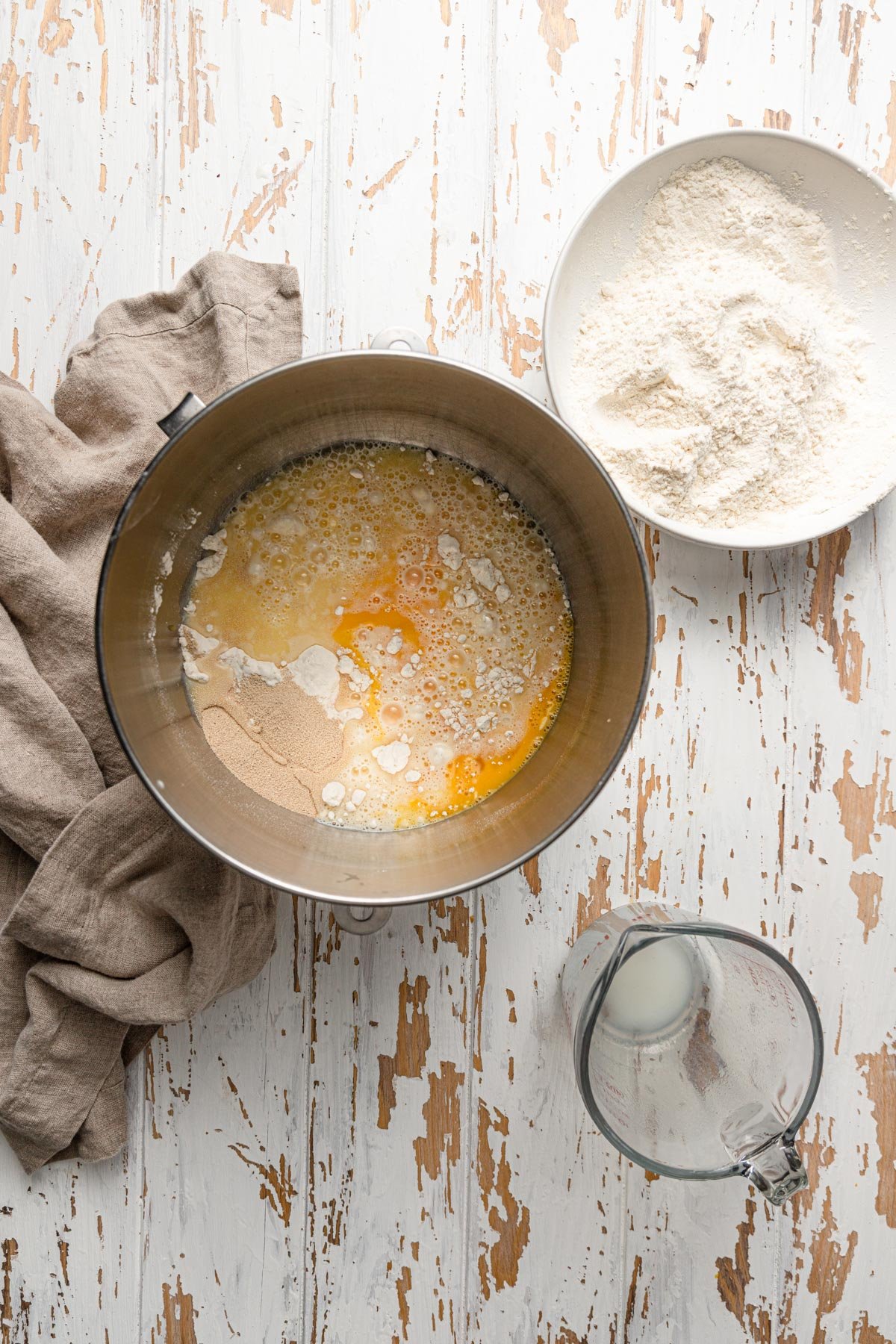 A mixer bowl with milk, yeast and eggs, next to a bowl with flour and a kitchen towel.