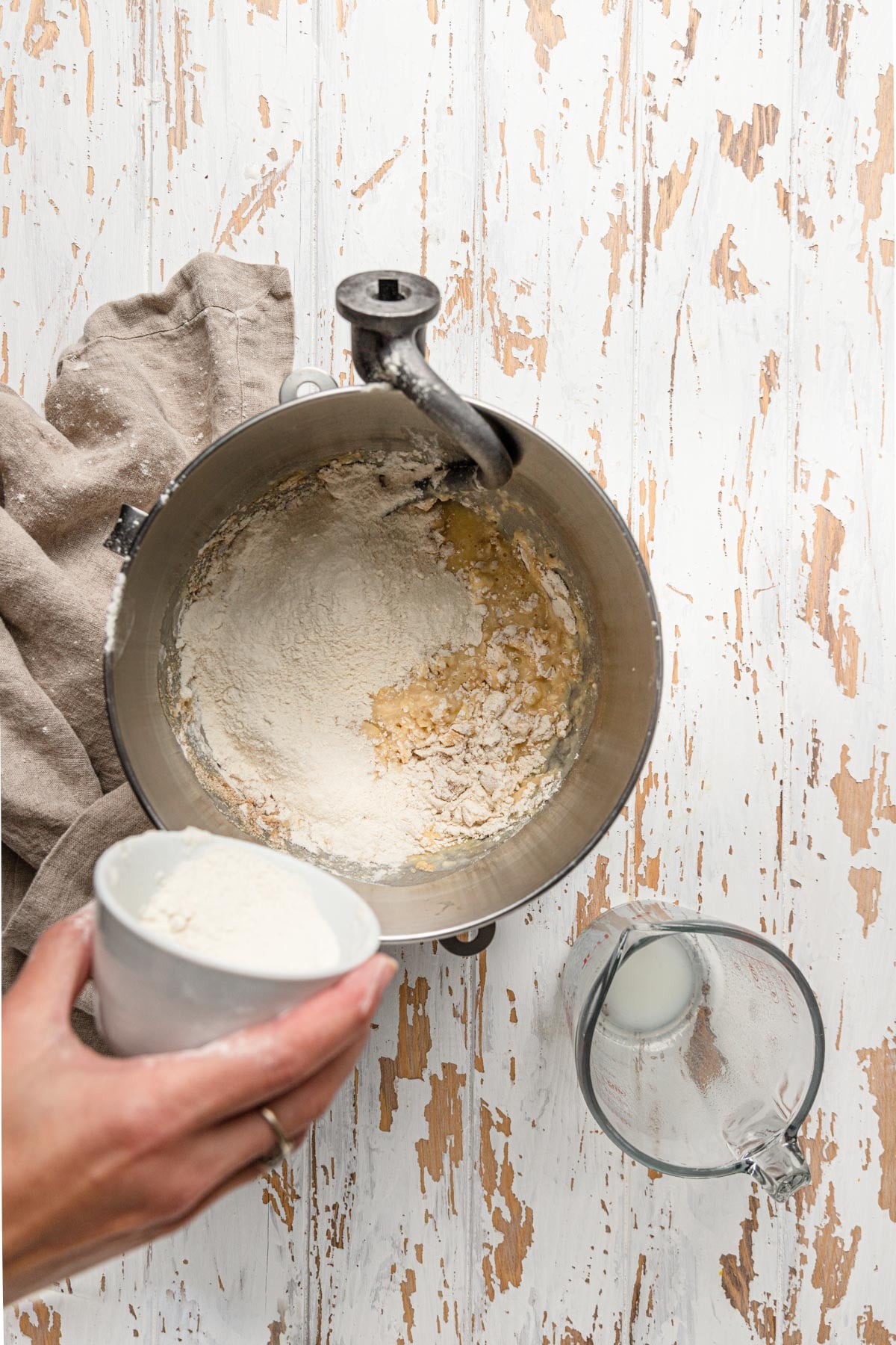 A mixer bowl with flour mixture and a hand adding flour. Dough hook in the mixer bowl.
