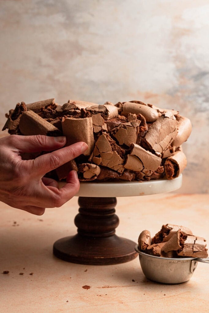 A hand placing pieces of chocolate meringue over the sides of concord cake.