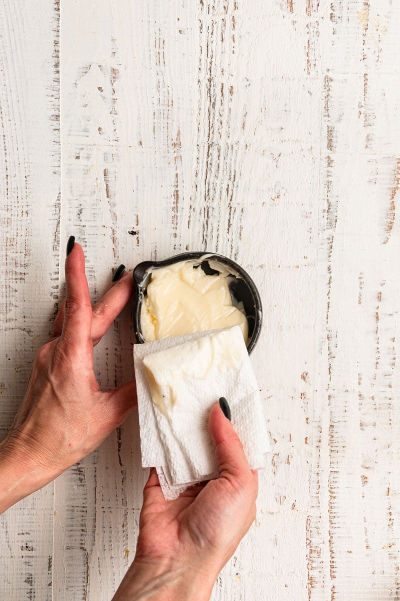 Hands dipping paper towel into a bowl with soft butter.