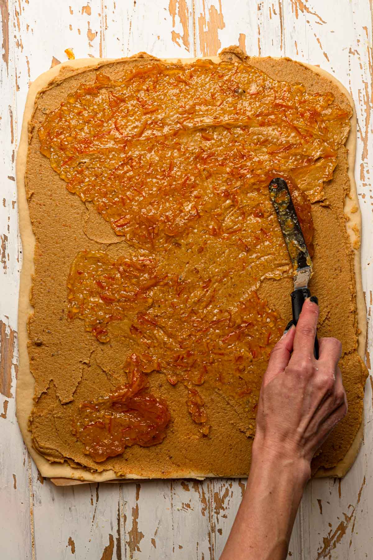 A hand spreading orange marmalade over rolled dough.