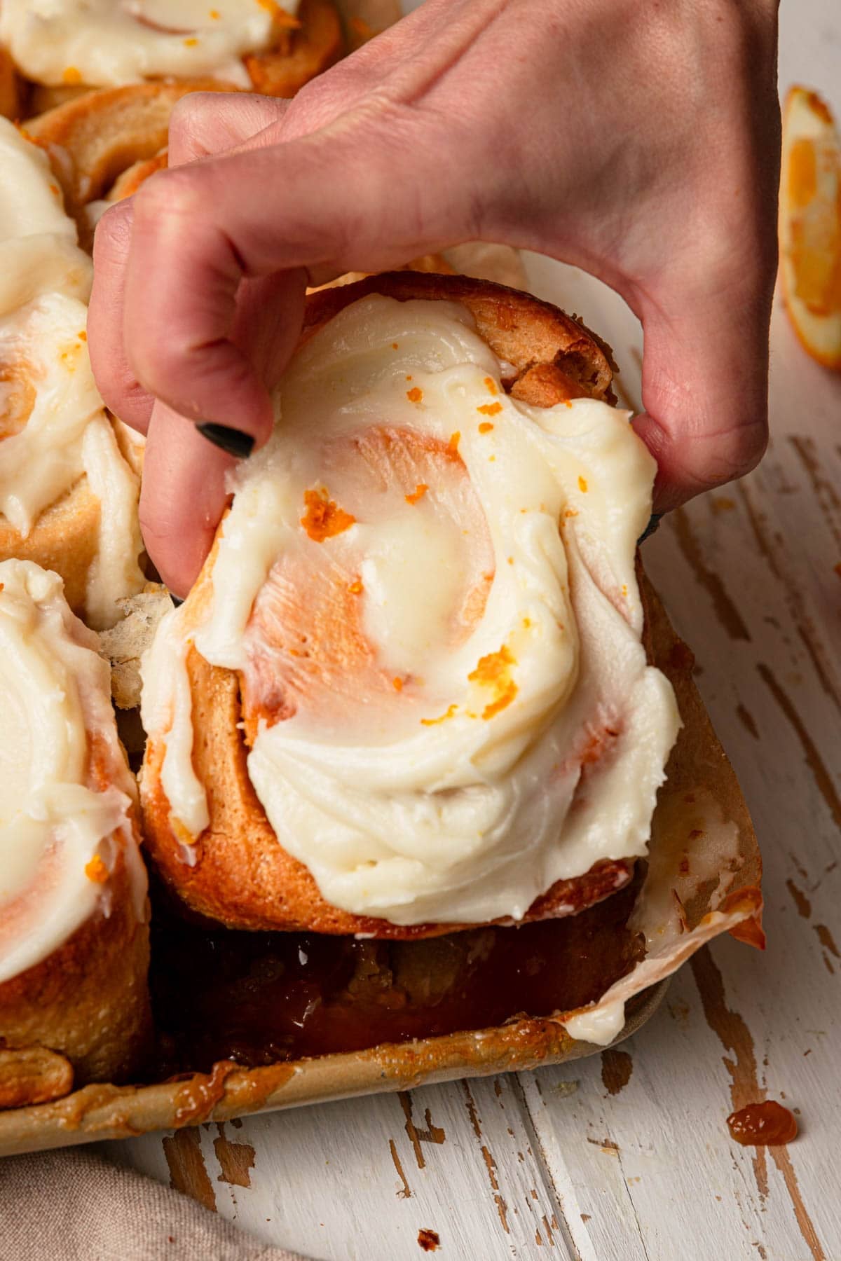 A hand lifting an orange roll topped with cream cheese frosting.