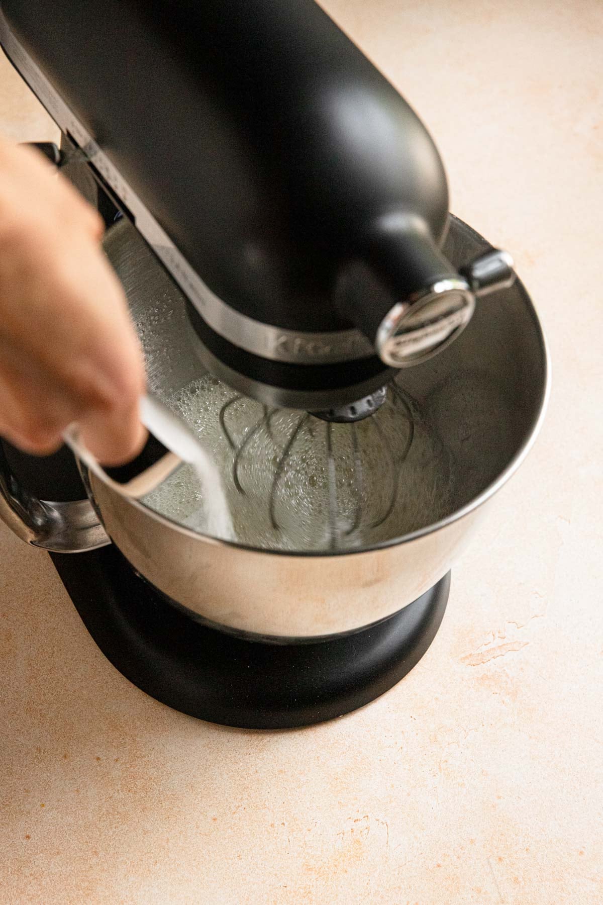 A hand pouring sugar into a stand mixer bowl with egg whites.