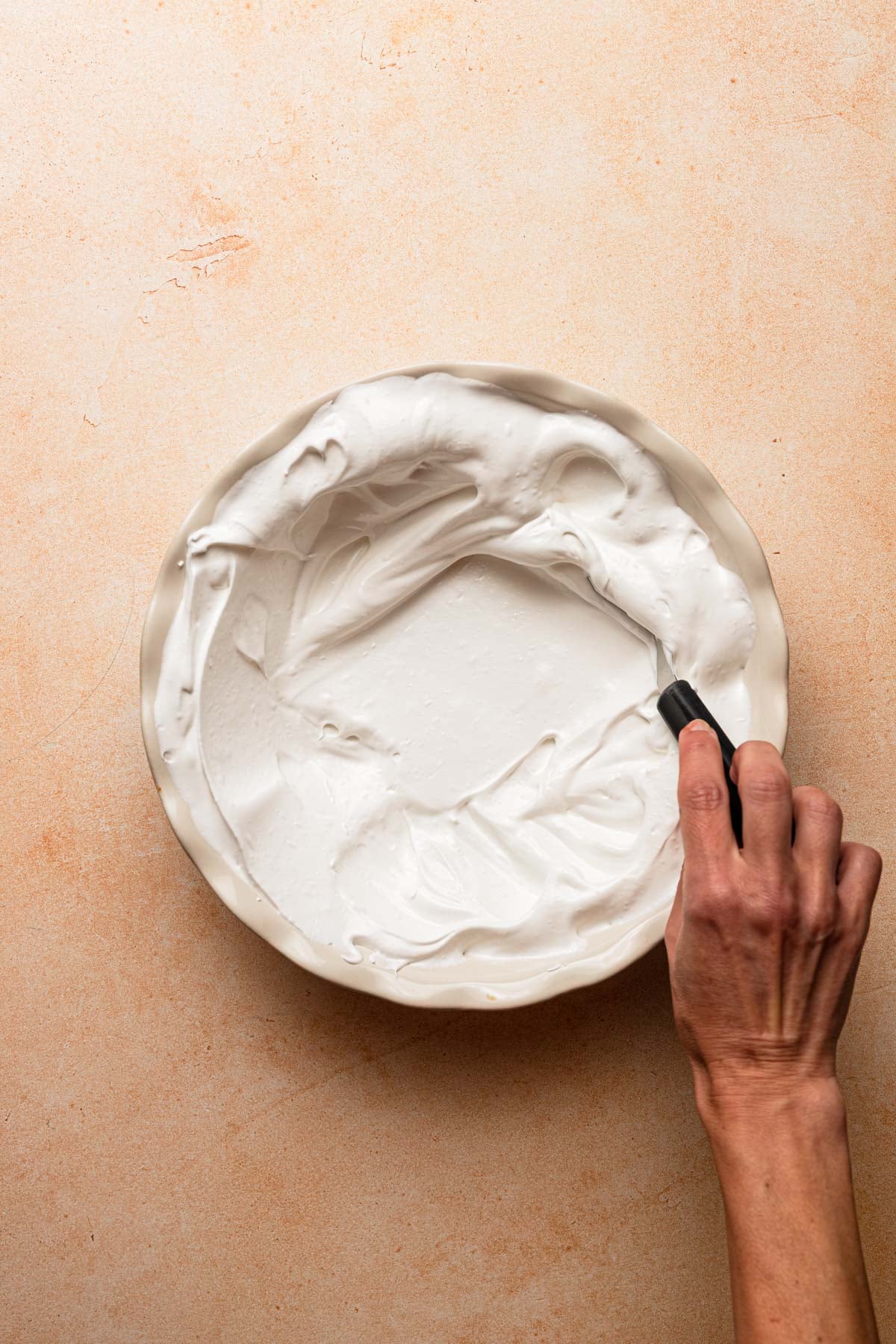 A hand spreading meringue onto a pie dish.