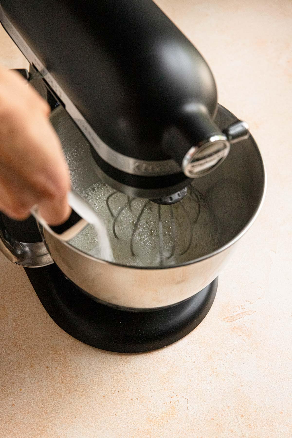 A hand pouring sugar to a bowl of a stand mixer with egg whites.