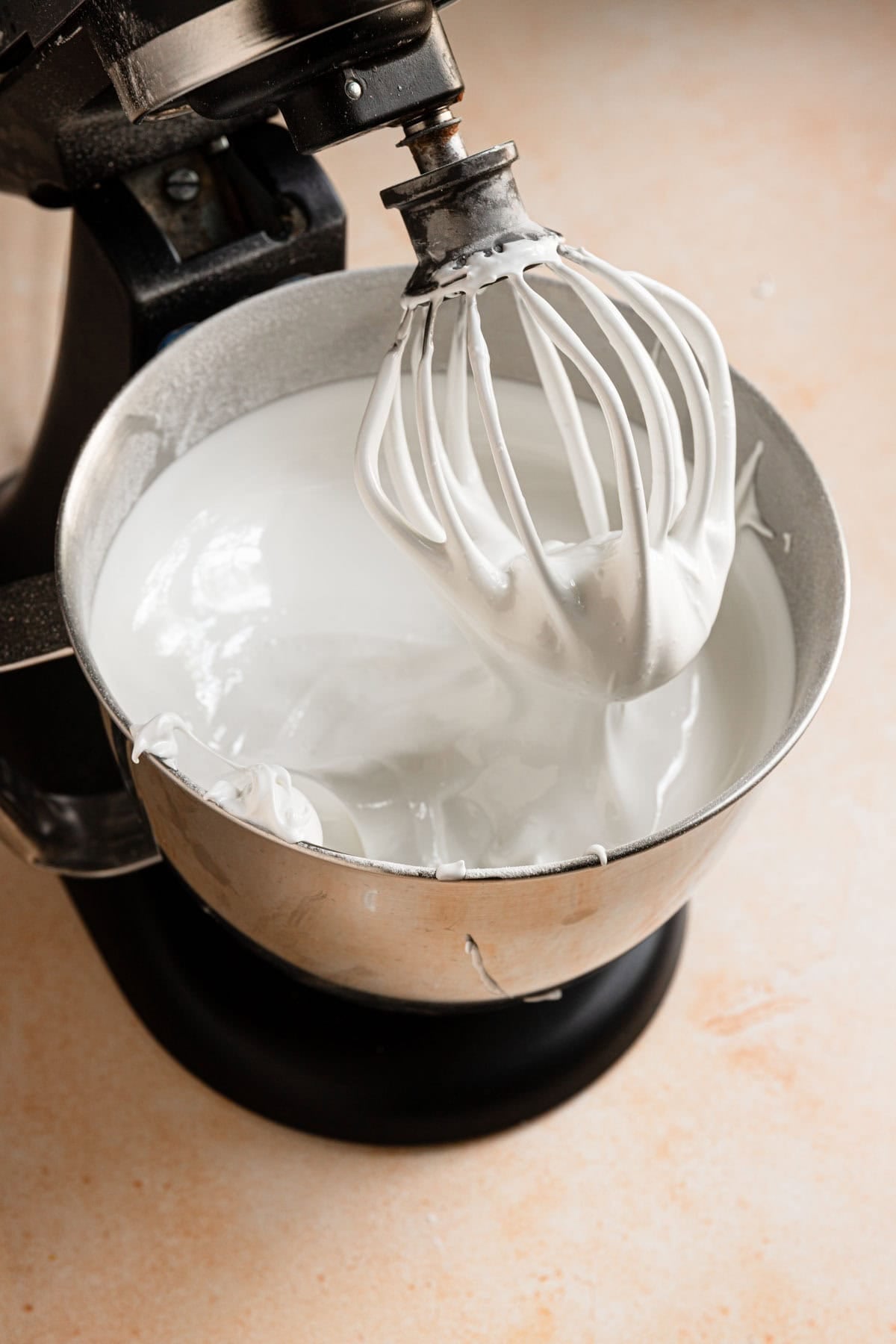 A stand mixer with a whisk attachment and a bowl full of meringue.
