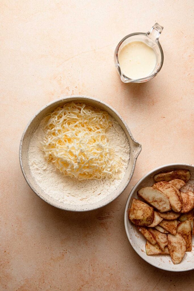 a bowl with flour and butter, next to a bowl with apple-cinnamon slices and a cup with buttermilk.